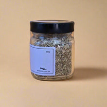 Load image into Gallery viewer, Glass jar with black lid containing dried herbs on a beige background